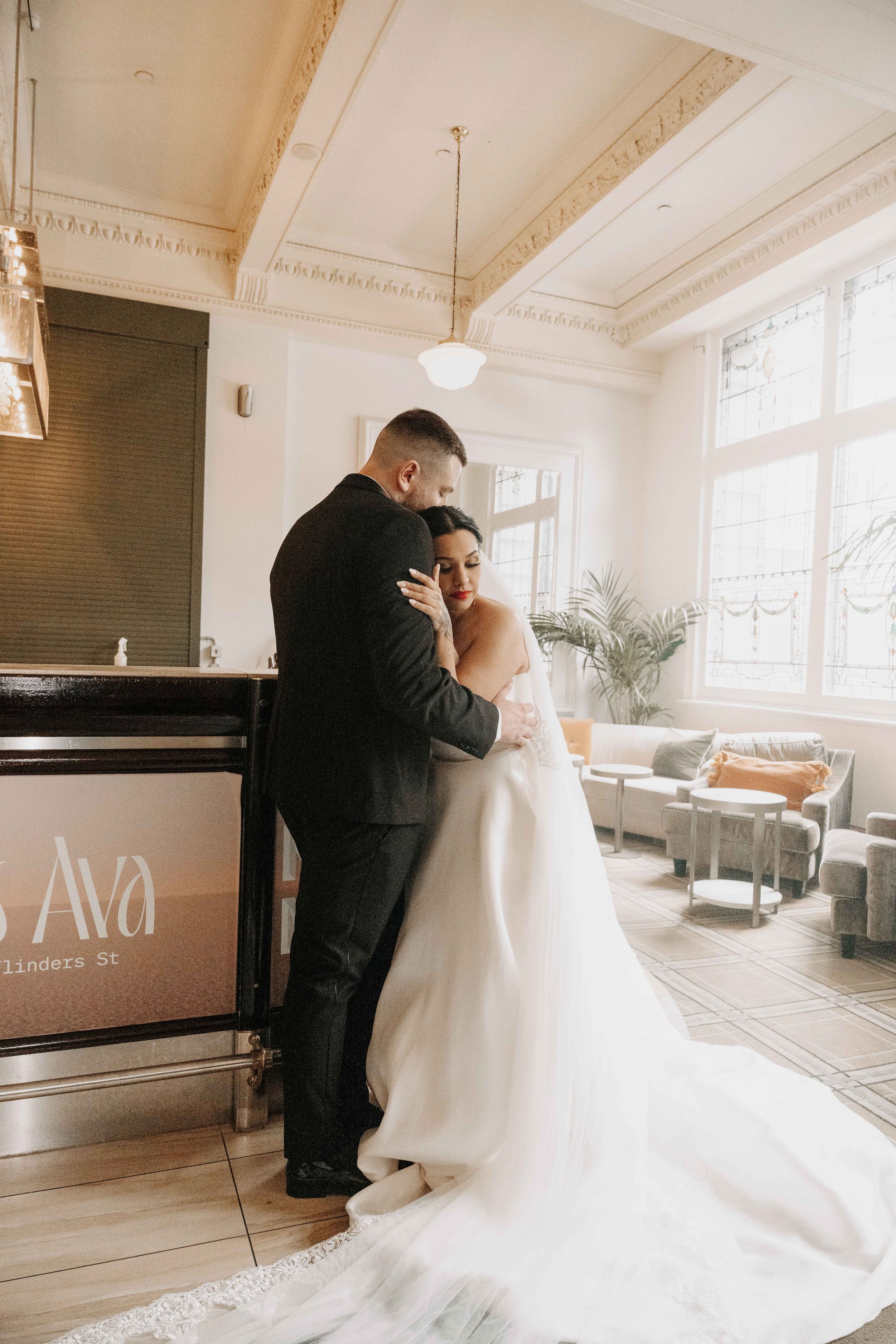 A bride and groom share an intimate embrace in an elegant venue with ornate ceilings and large windows on their wedding day.