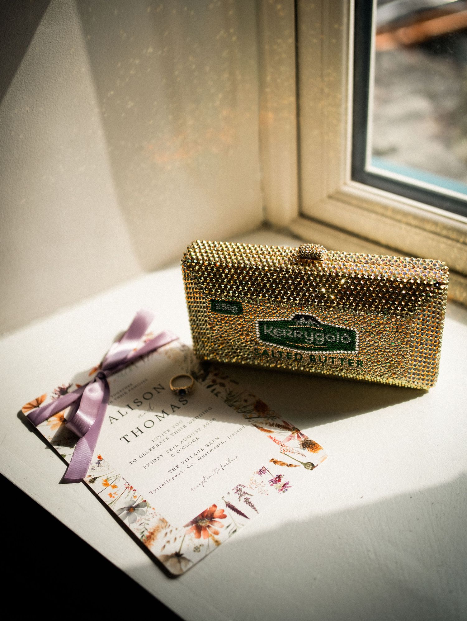 Gold rhinestone Kerrygold clutch purse on windowsill with floral wedding invitation and ring.
