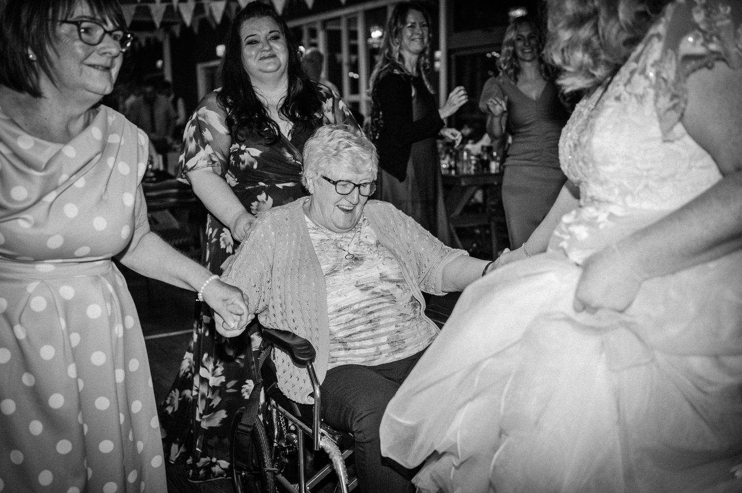 Black and white photo of bride dancing with elderly woman in wheelchair at wedding reception.