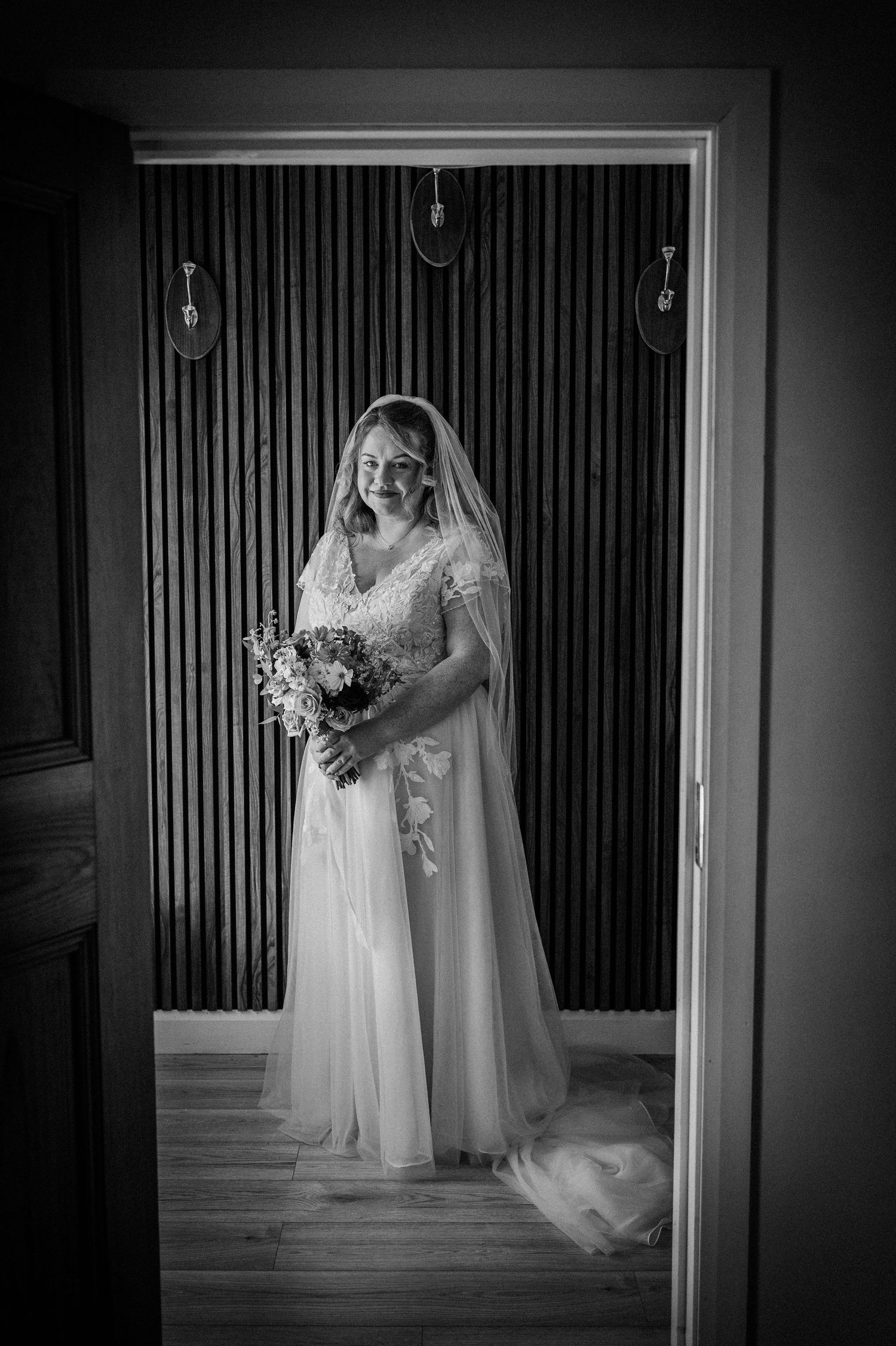 Black and white photo of a smiling bride in a lace gown and veil holding a bouquet, framed in a doorway.