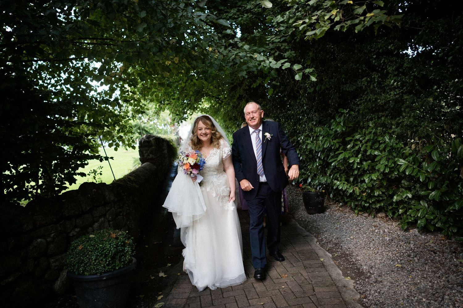 Bride in white gown and groom in suit walk together along a garden path surrounded by lush green trees.