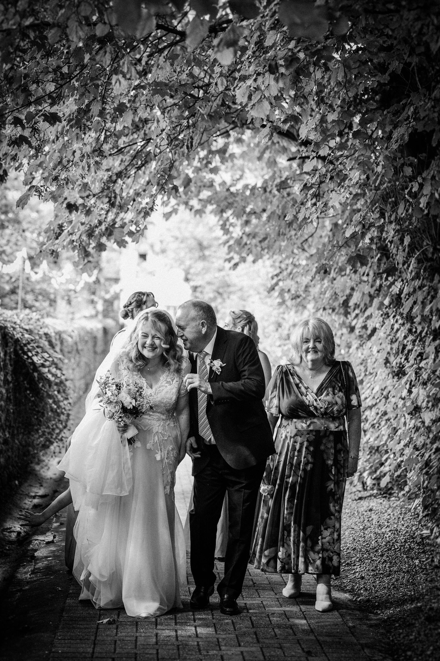 Bride in white gown walks with family under a leafy archway in black and white wedding photo.