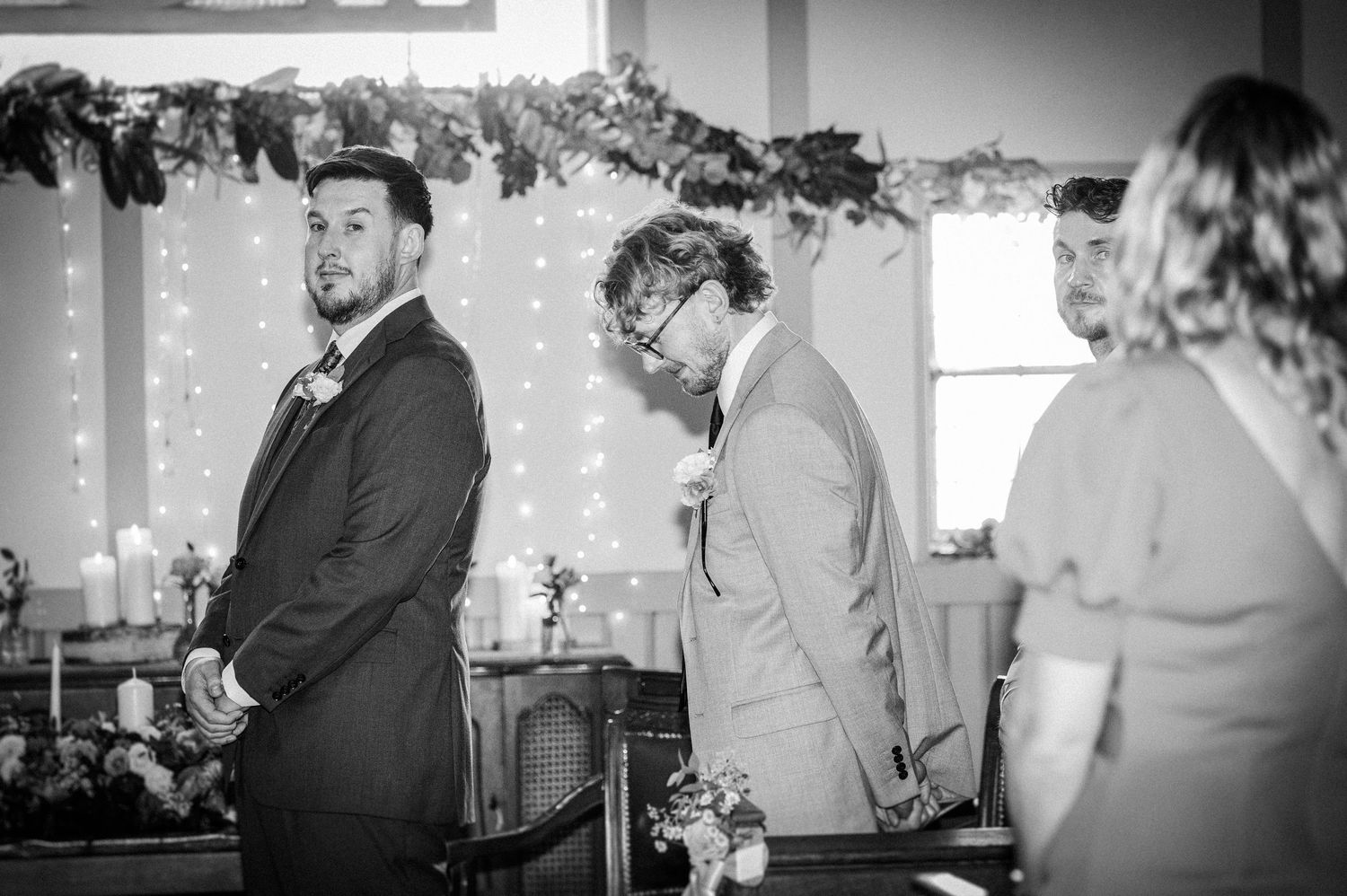 Black and white wedding ceremony photo of groomsmen standing at the altar with fairy lights and floral decorations.
