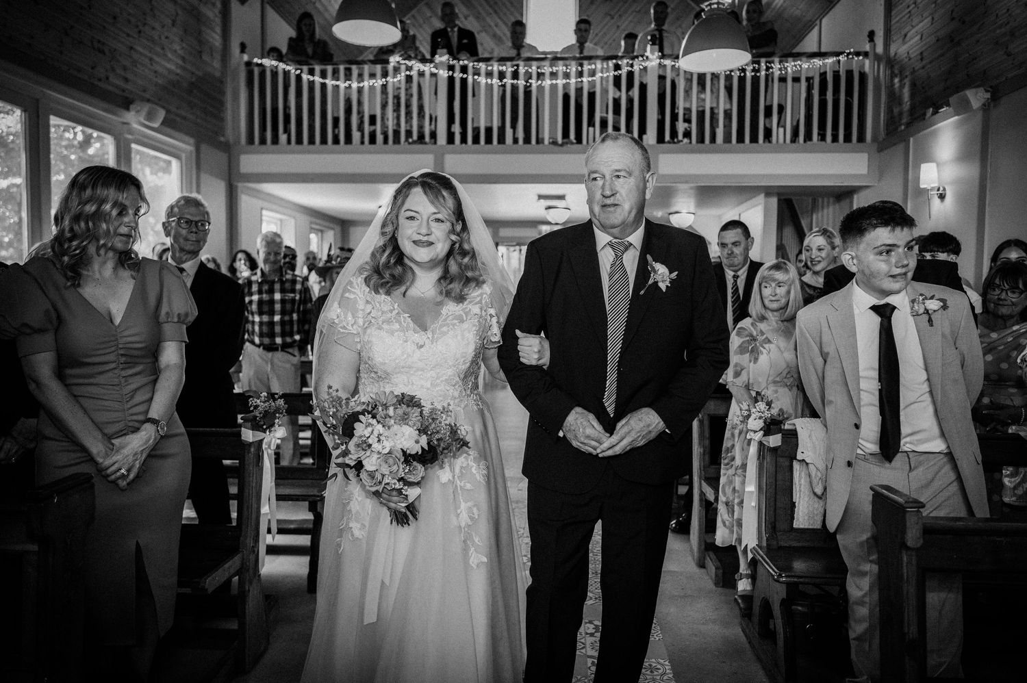 Bride walks down the aisle with escort in black and white wedding ceremony photo.