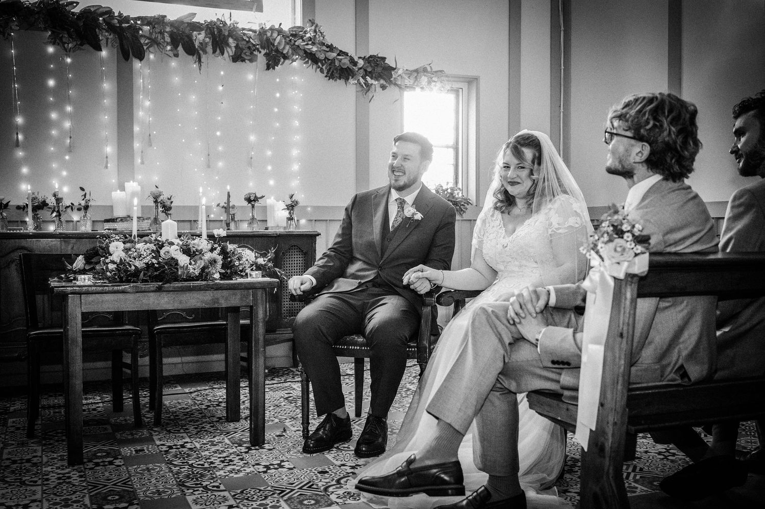 Black and white photo of a bride and groom holding hands during an intimate wedding ceremony indoors.