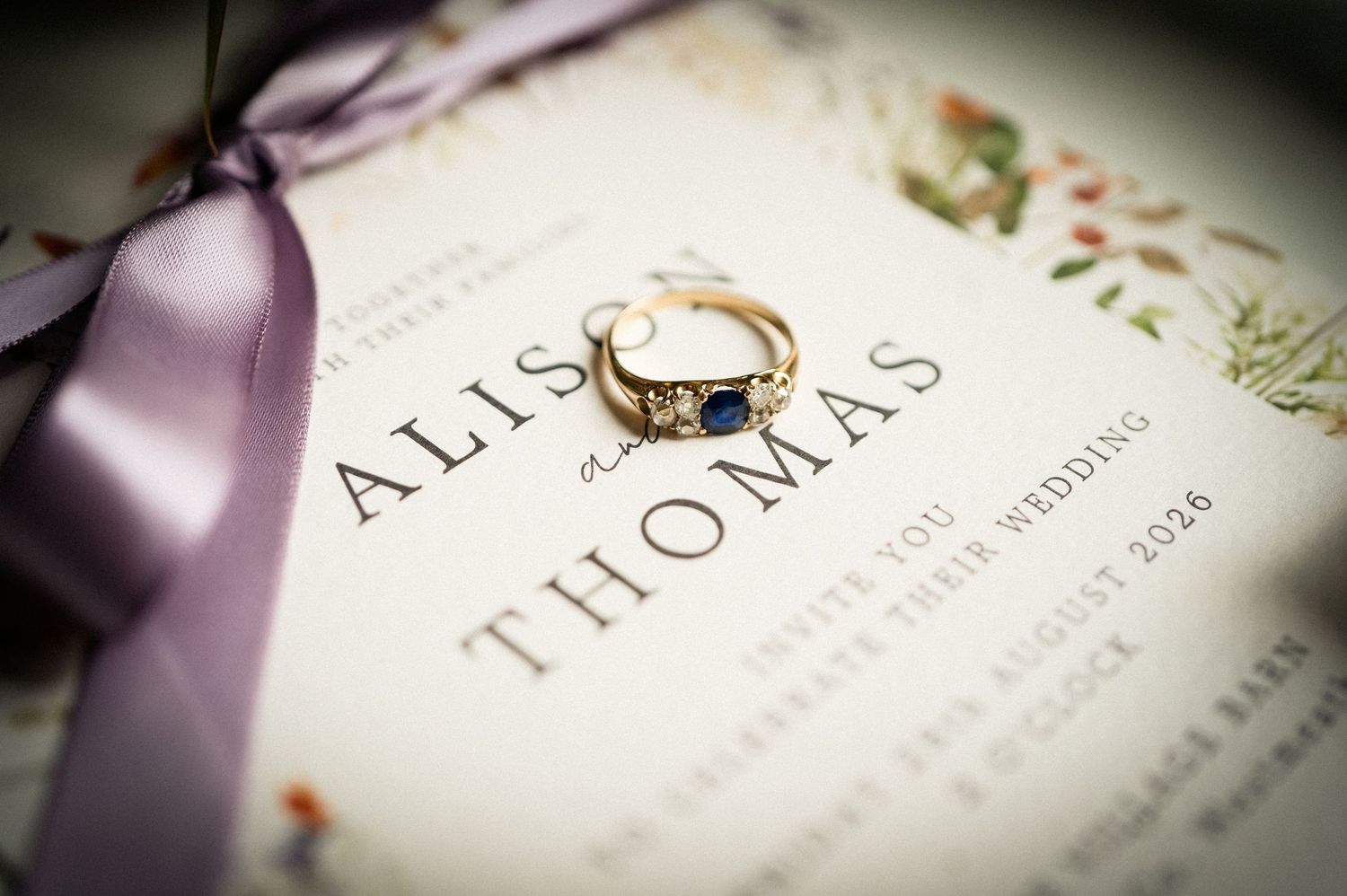 Gold engagement ring with blue sapphire on Alisa and Thomas wedding invitation with purple ribbon.