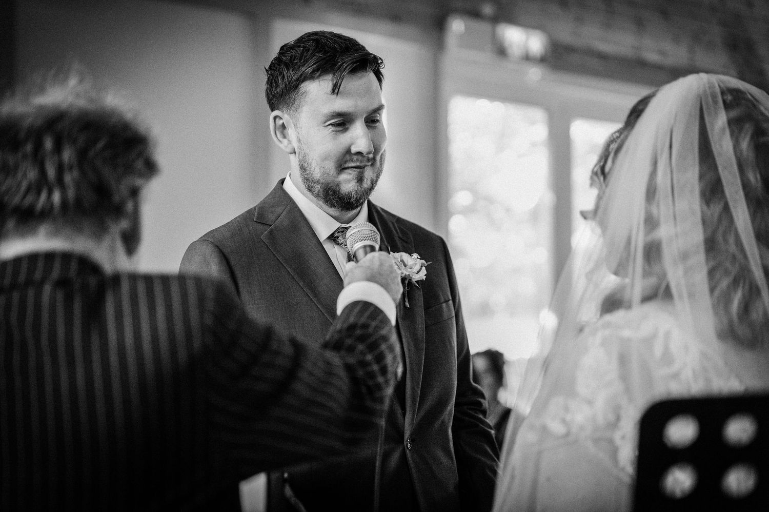 Black and white wedding photo of groom smiling as bride adjusts his boutonniere during ceremony.