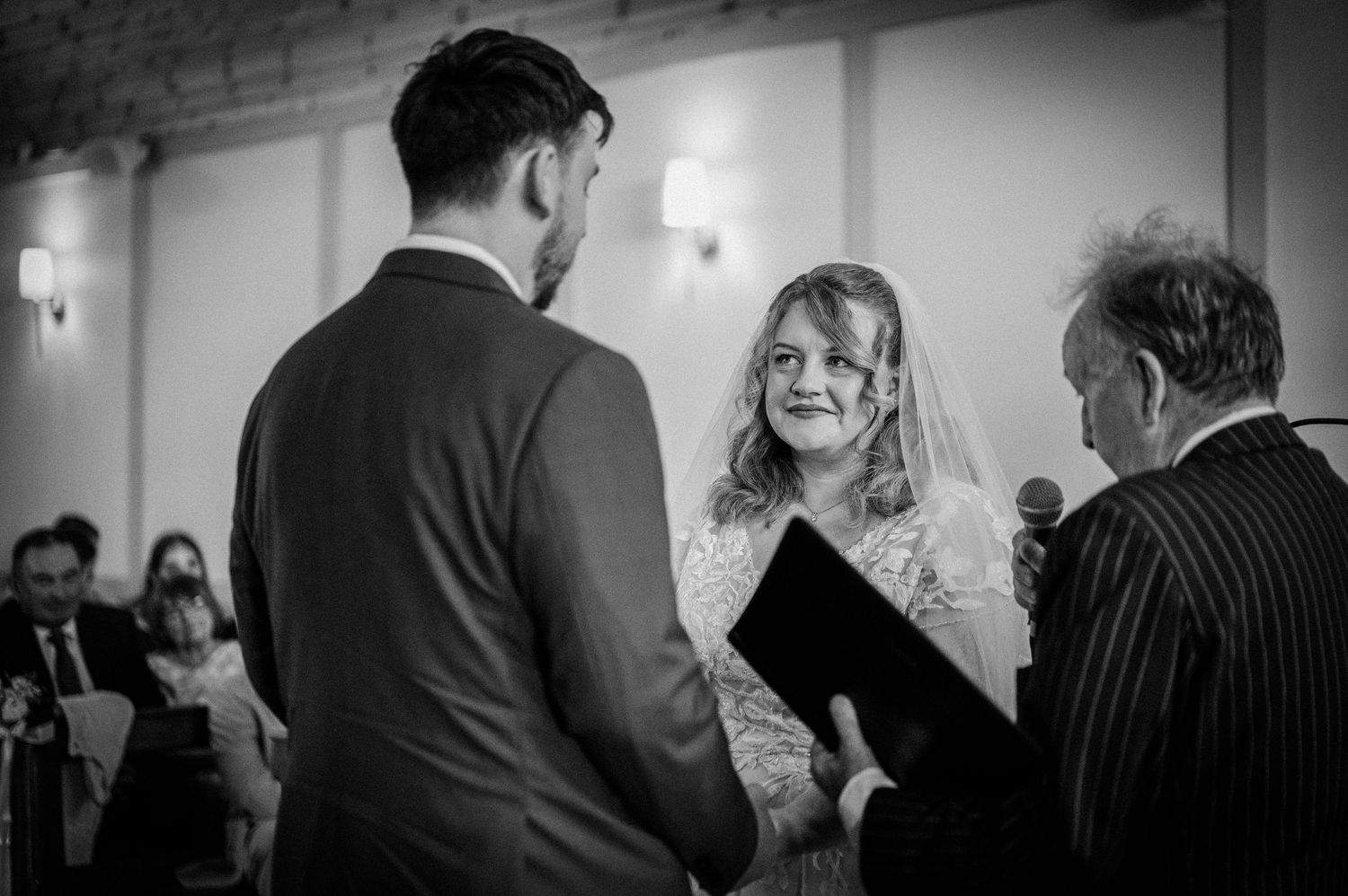 Bride and groom exchange vows before officiant in elegant barn wedding ceremony, black and white.