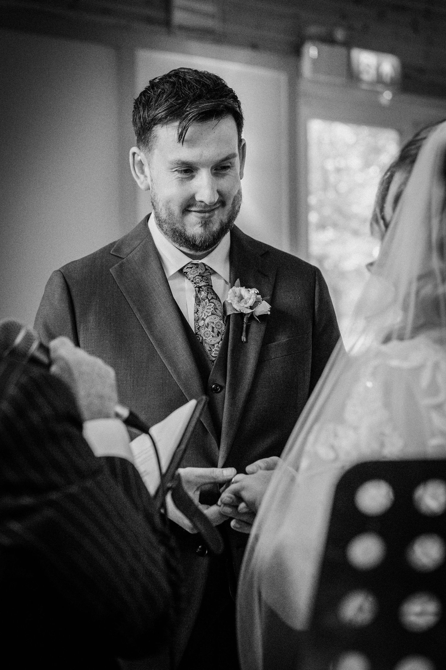 Groom smiling warmly during wedding ring exchange ceremony, wearing suit with floral boutonniere.