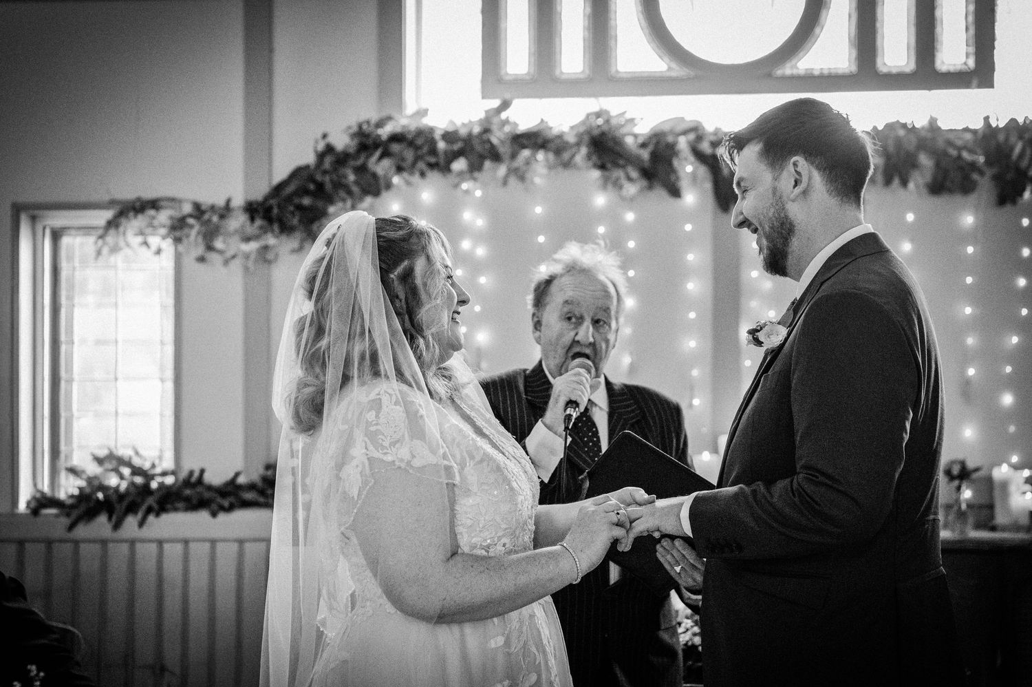 Black and white wedding ceremony photo of bride and groom exchanging vows with officiant and fairy lights backdrop.