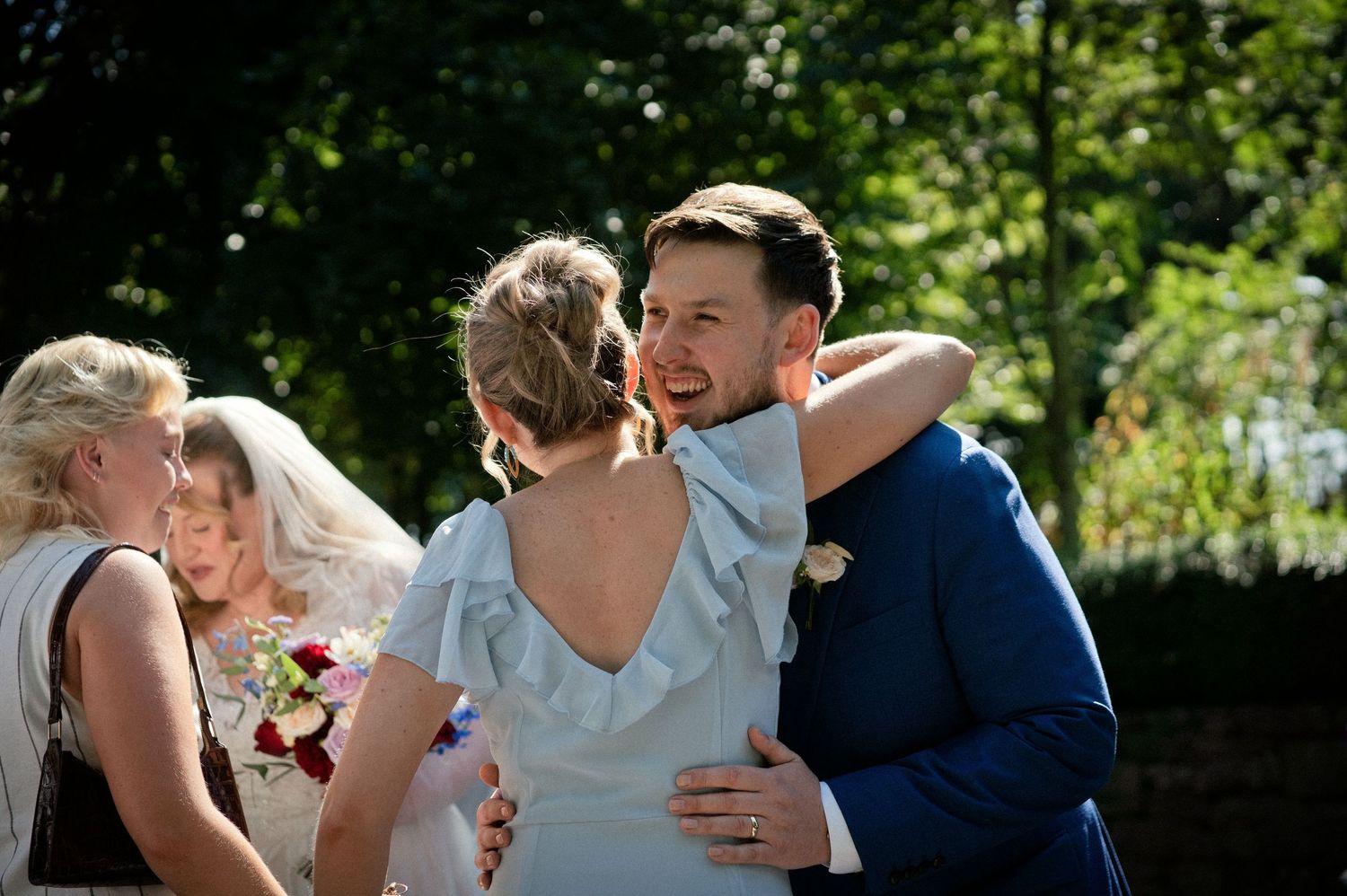 Groom in navy suit embracing bride in backless blue dress outdoors at wedding celebration.