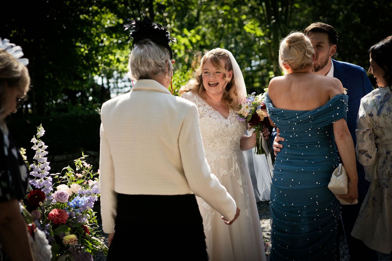 Joyful bride in lace gown greeting guests outdoors while holding a colorful bouquet at her wedding.