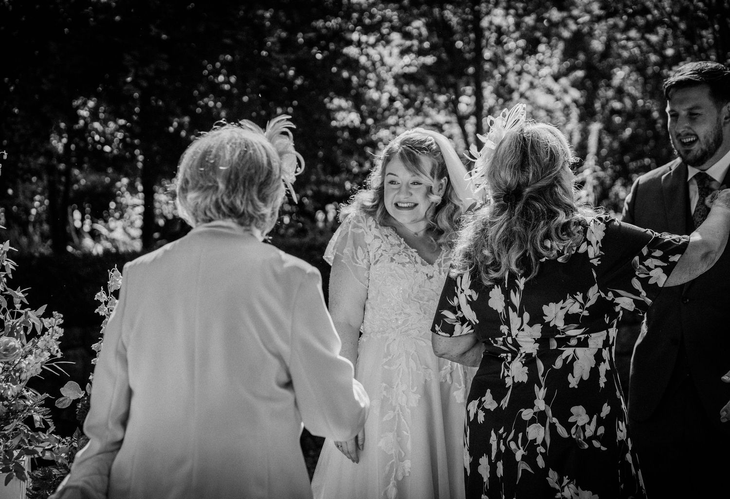 Black and white wedding photo of a smiling bride in lace gown greeting guests outdoors among trees.