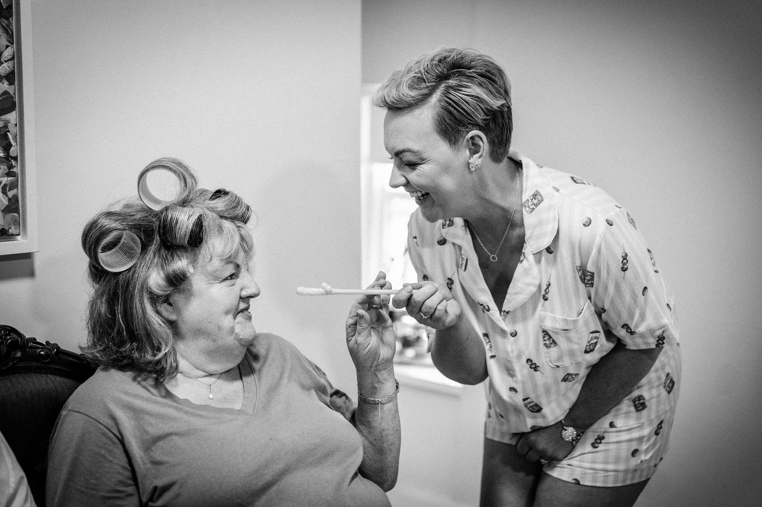 Black and white photo of two women laughing together, one in hair rollers, sharing a joyful moment during wedding preparations.