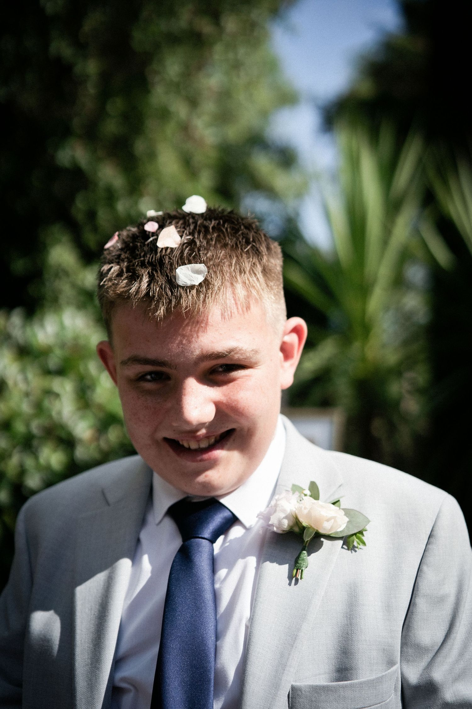 Young boy in gray suit with blue tie and boutonniere smiles with flower petals in his hair at a wedding.