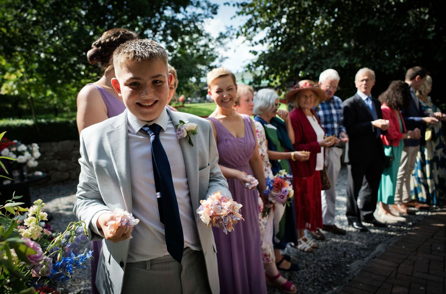 Young boy in gray suit holding flower petals at an outdoor wedding reception with guests lined up behind him.
