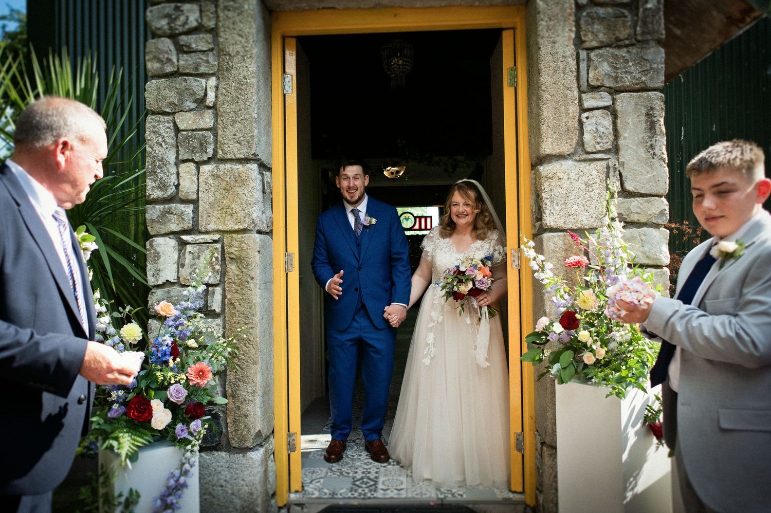 Newlywed couple exits stone church doorway holding hands as guests toss flower petals in celebration.