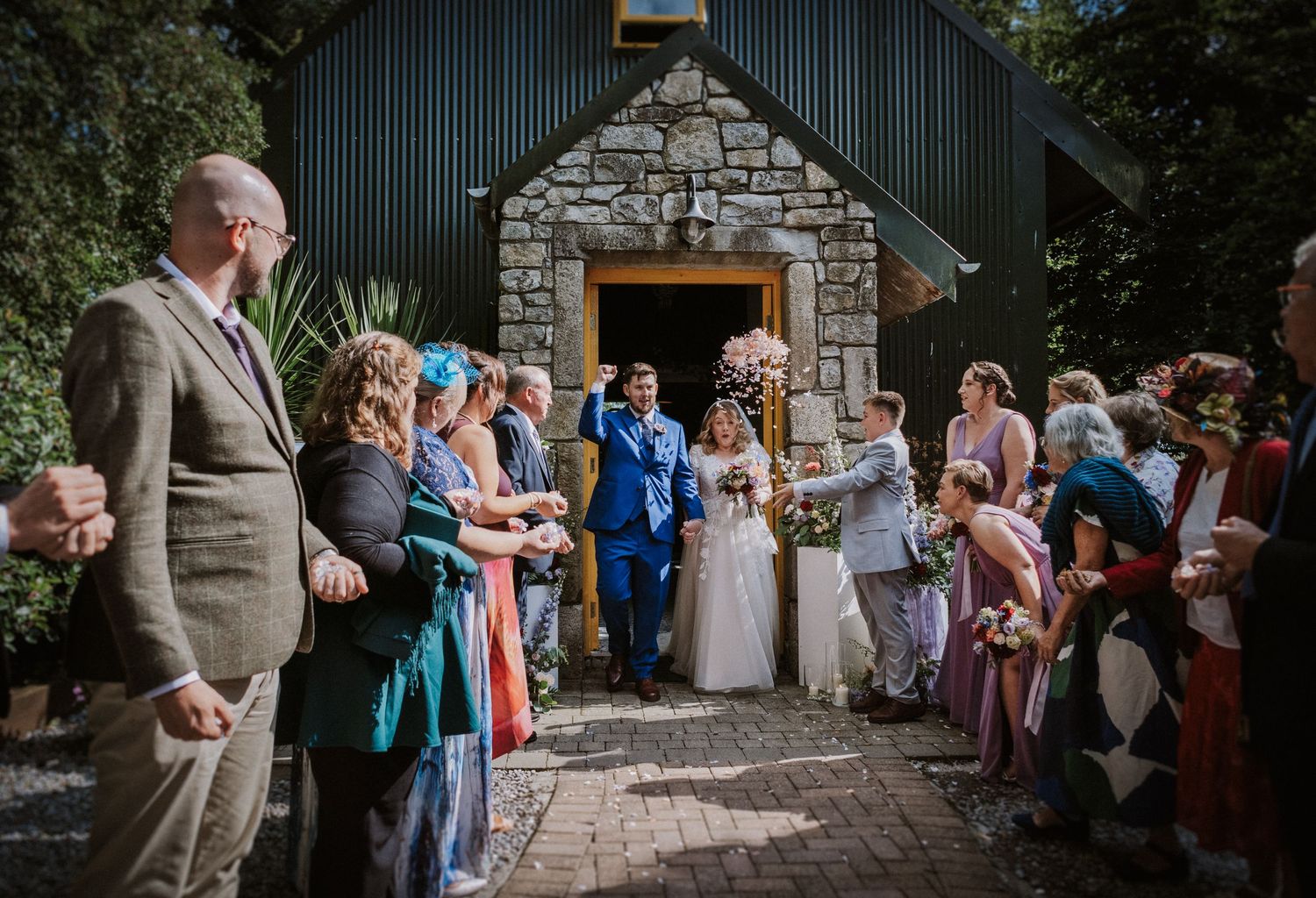 Bride and groom exit stone chapel greeted by cheering wedding guests on sunny day.