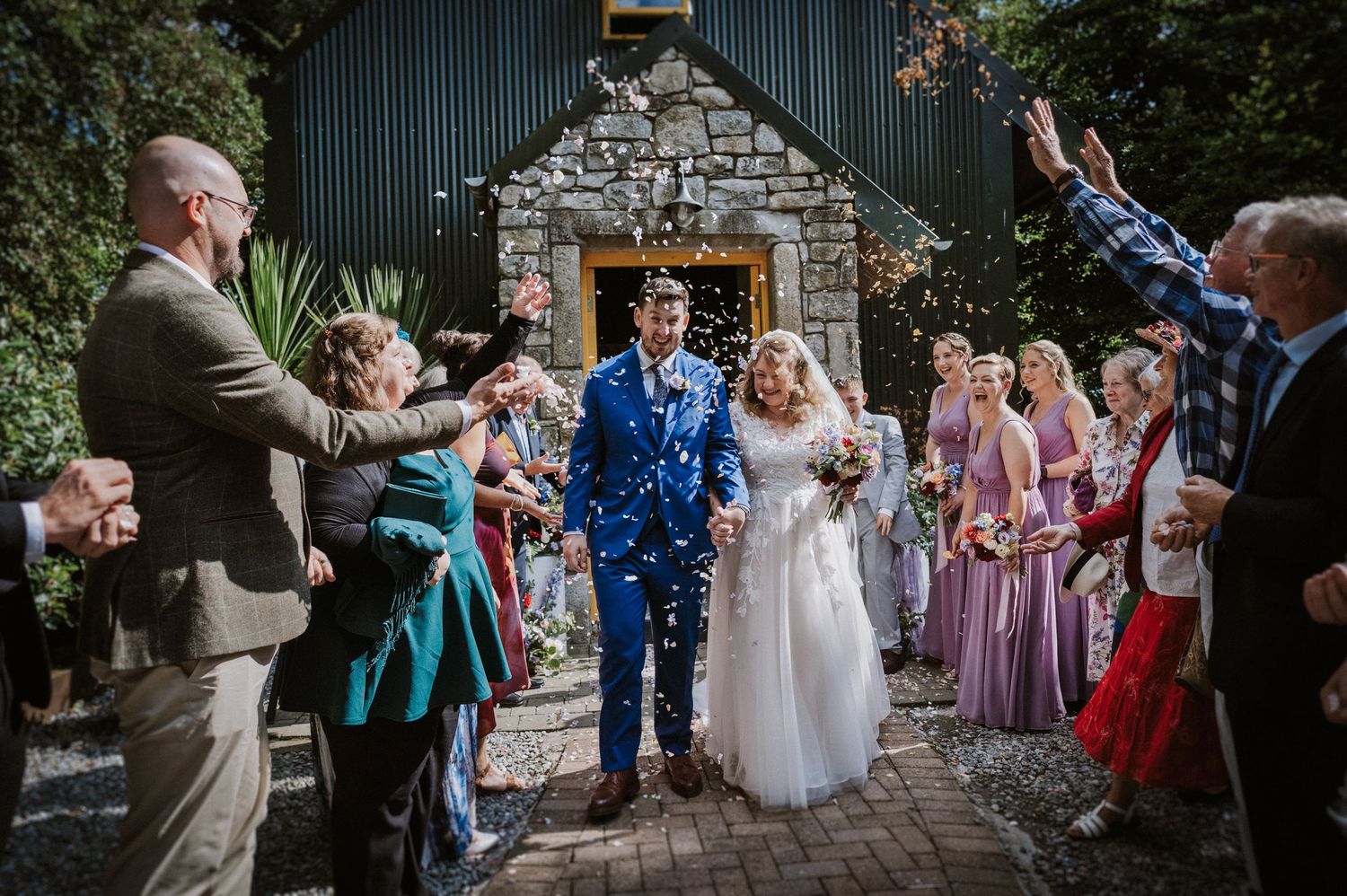 Bride and groom exit stone church as wedding guests shower them with colorful confetti outdoors.