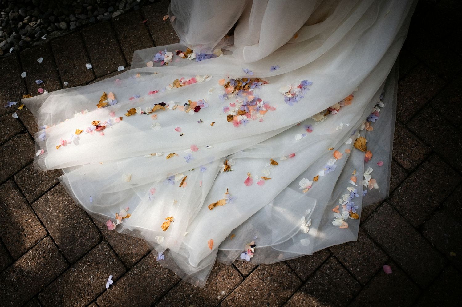 Wedding dress train covered in colorful flower confetti petals on brick floor.
