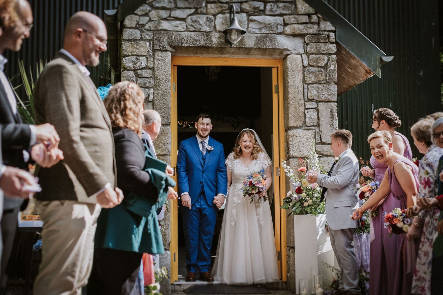 Bride and groom exit stone chapel as guests celebrate with confetti outside.