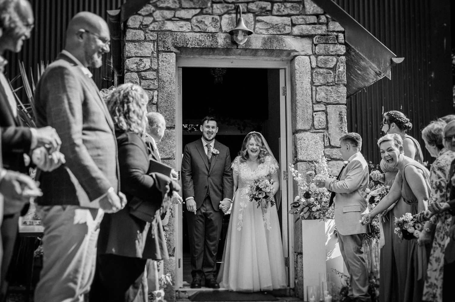 Bride and groom exit stone church doorway as wedding guests celebrate with confetti.