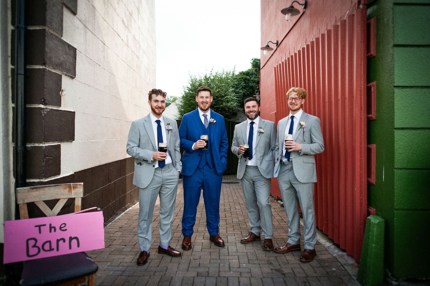 Groom in blue suit stands with three groomsmen in grey suits holding drinks outside a barn venue.