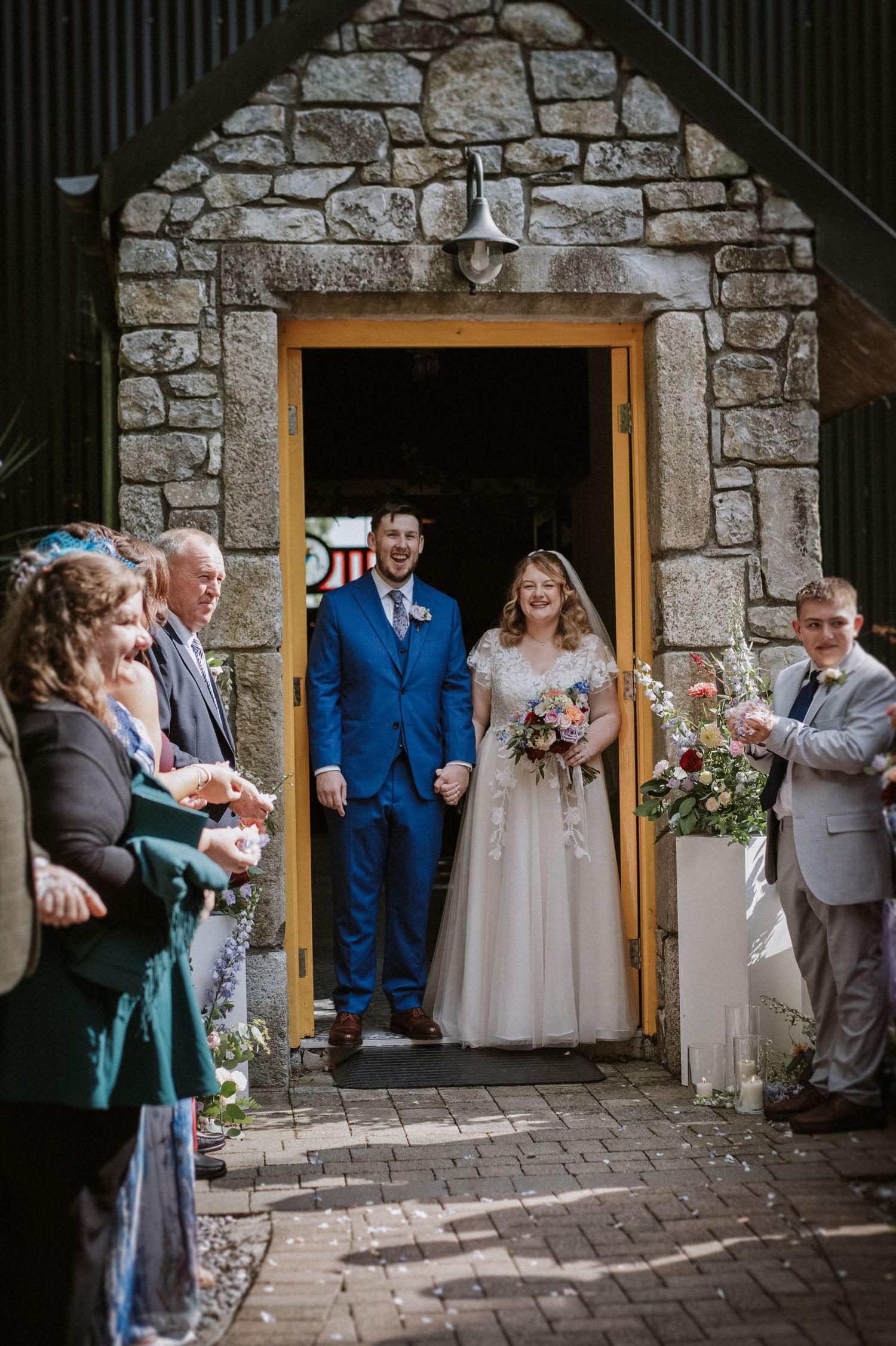 Smiling bride and groom in blue suit exit rustic stone venue to cheering guests.