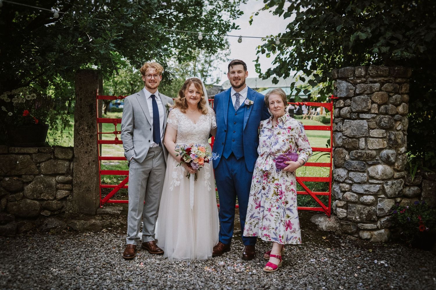 Wedding party of four poses by red gate and stone wall, bride in tulle gown with groom in blue suit.