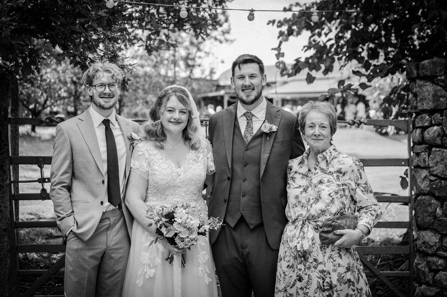 Black and white wedding photo of bride and groom posing with two guests outdoors at a rustic venue.