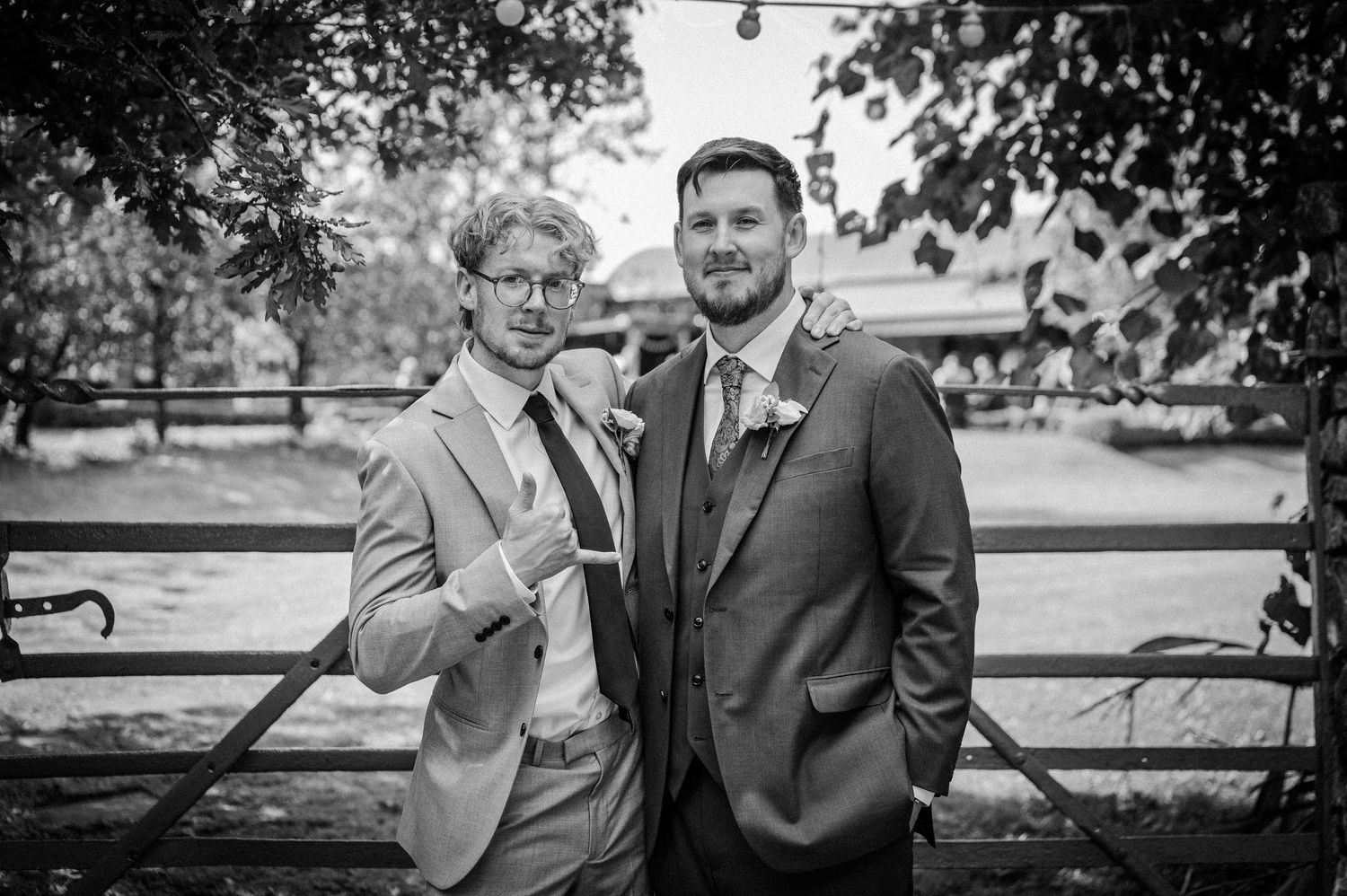 Two men posing together outdoors at a wedding, one in a light suit and one in a dark suit.