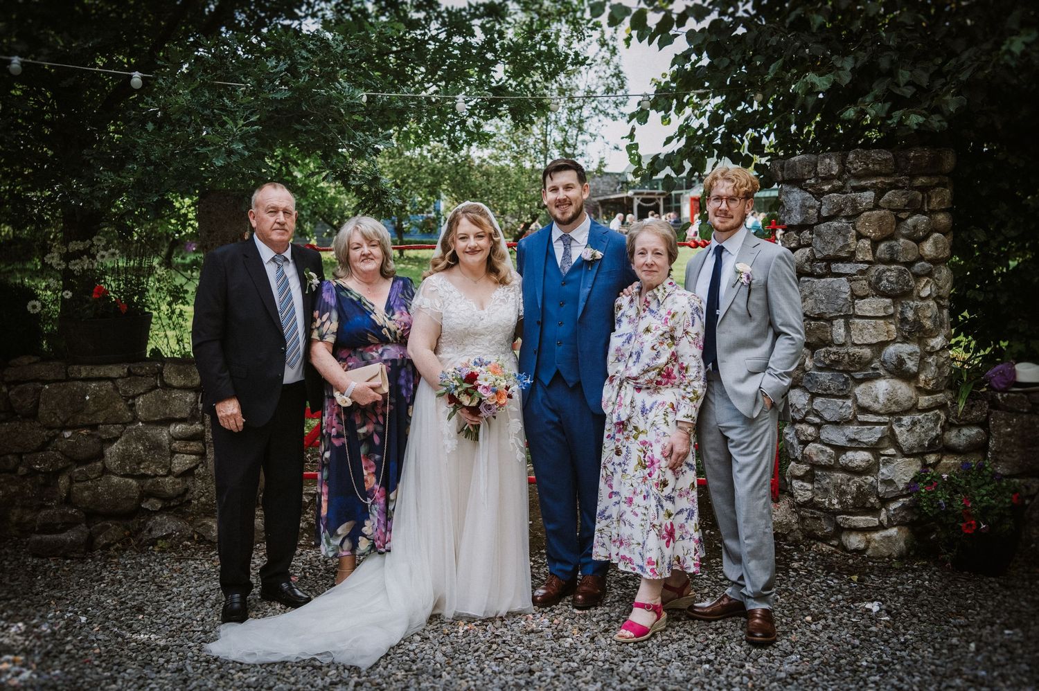 Wedding party of six poses outdoors against a stone wall with bride in lace gown and groom in blue suit.