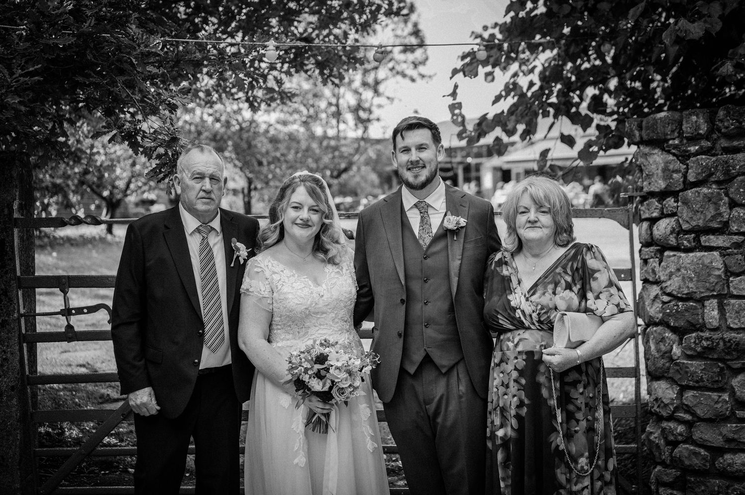 Bride and groom pose with family members in formal wedding attire near a stone wall in black and white.