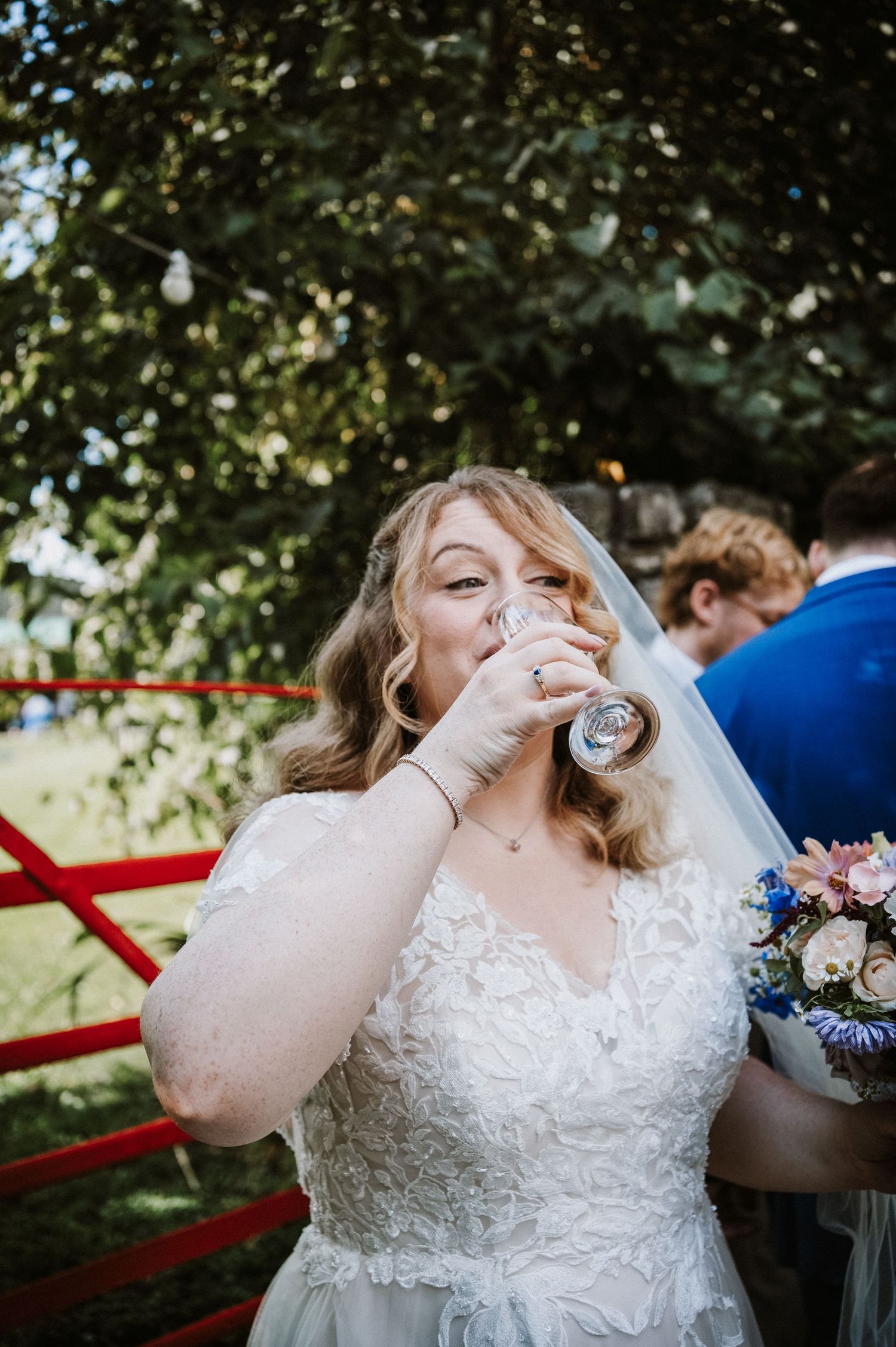 Bride in lace wedding dress and veil drinking champagne while holding a floral bouquet outdoors.