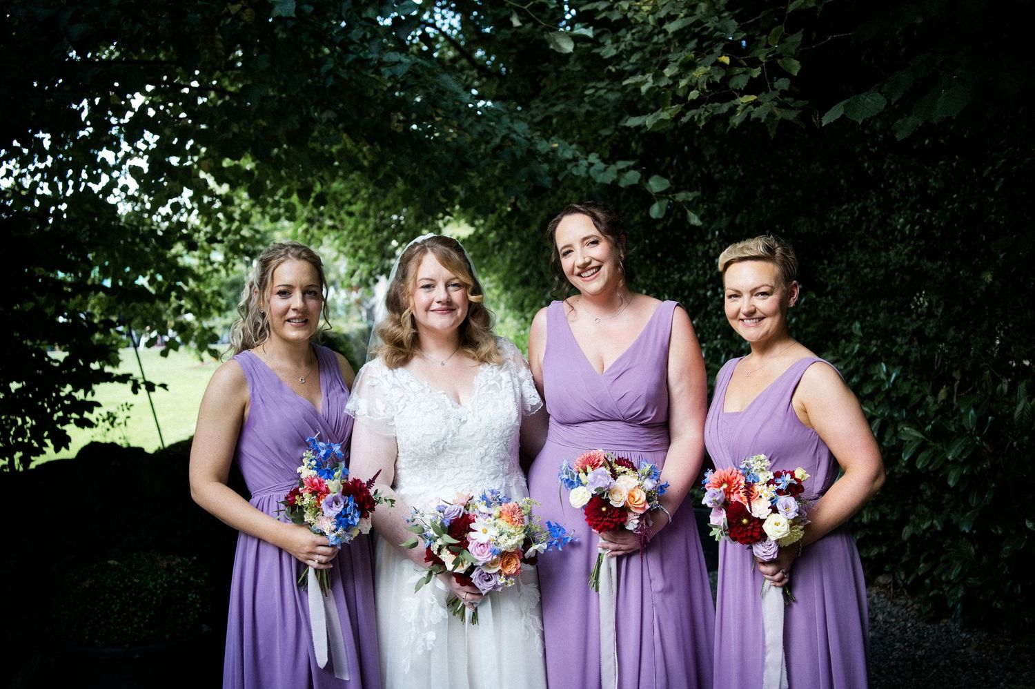 Bride with three bridesmaids in lavender dresses holding colorful bouquets outdoors.