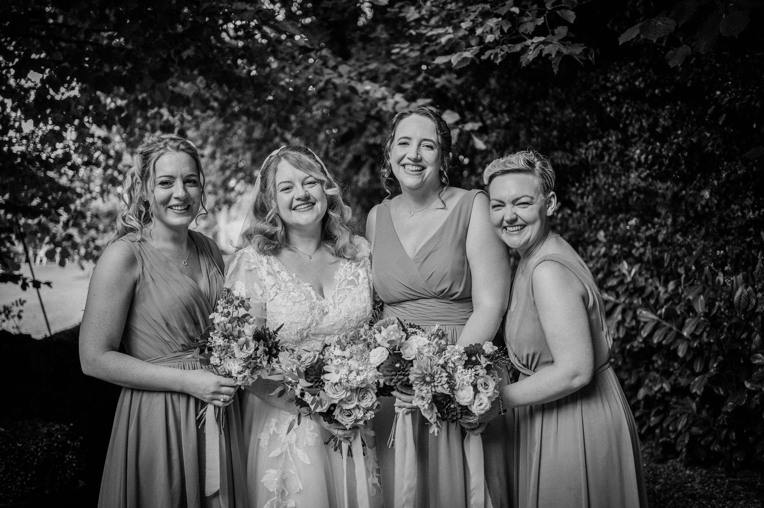 Black and white photo of a bride and three bridesmaids holding bouquets, smiling outdoors.
