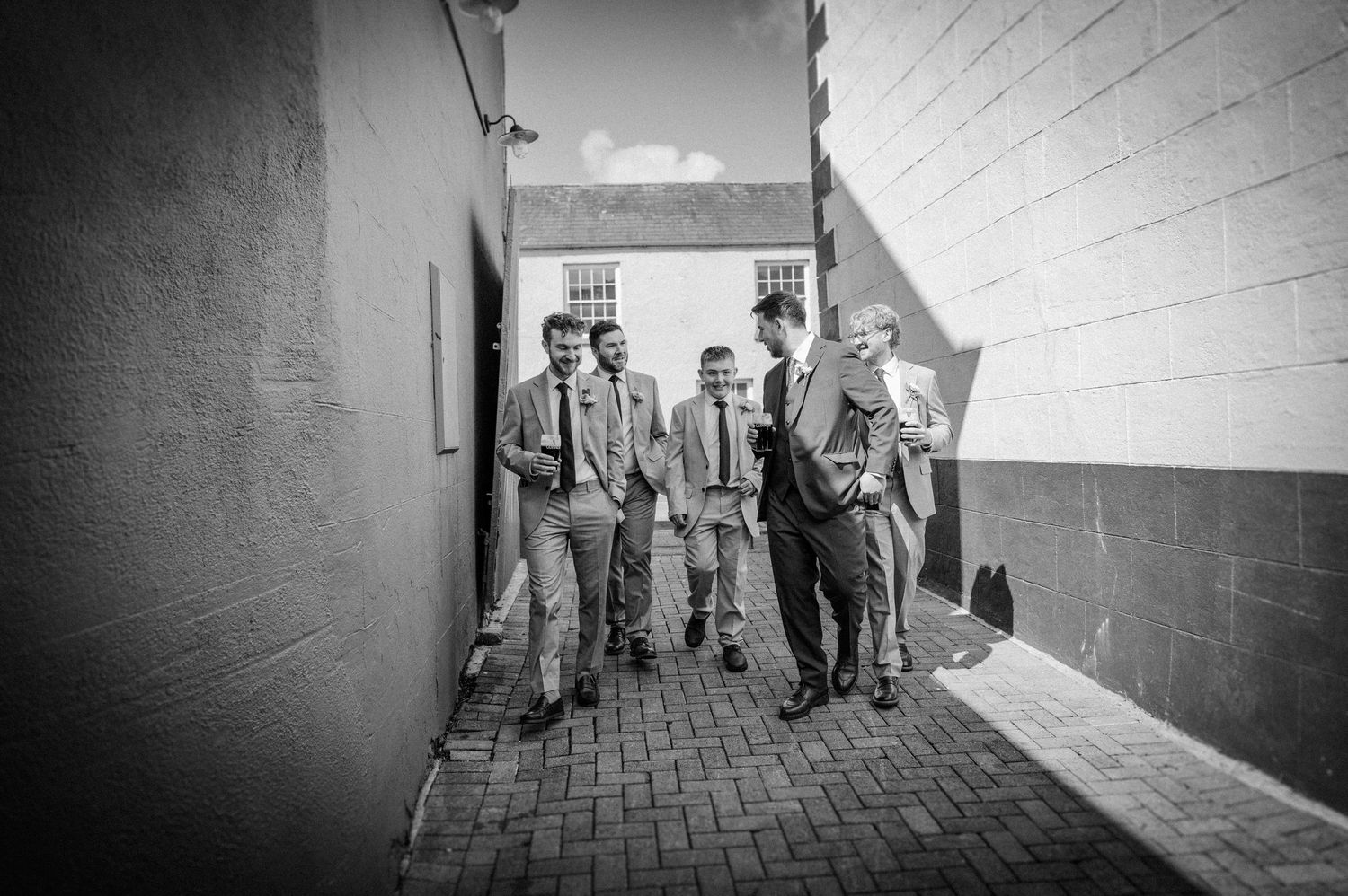Black and white photo of groomsmen in suits walking together through a narrow alleyway on a wedding day.