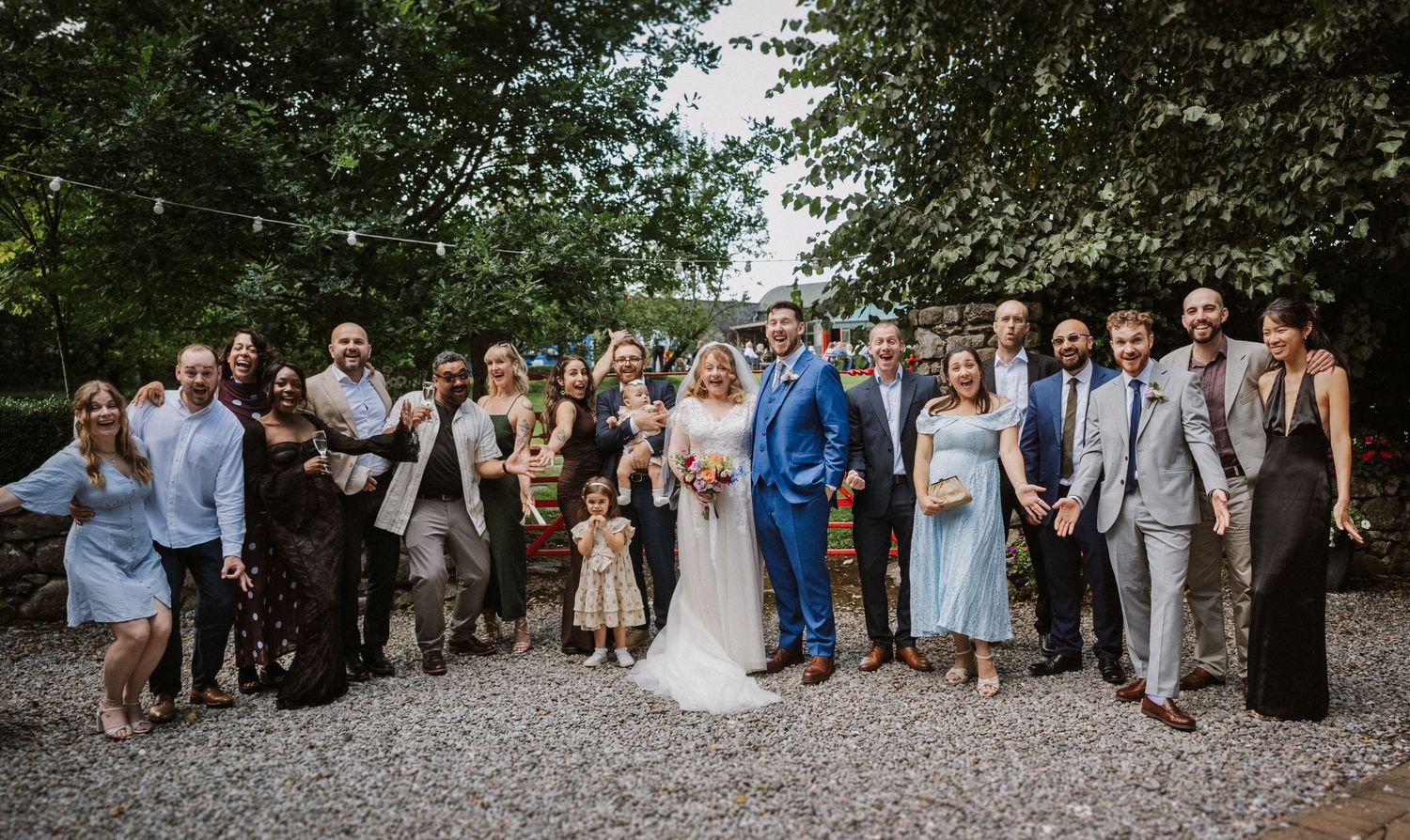 Large wedding party group photo with bride and groom surrounded by guests on a gravel path.