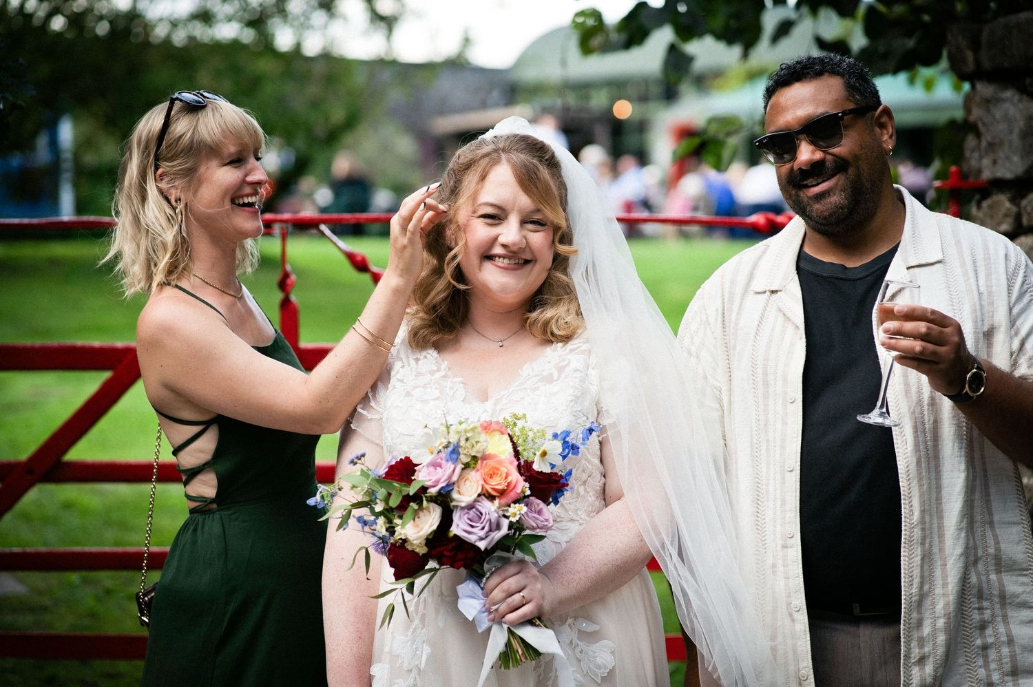 Bride holding colorful bouquet laughs with two guests adjusting her veil at outdoor wedding reception.