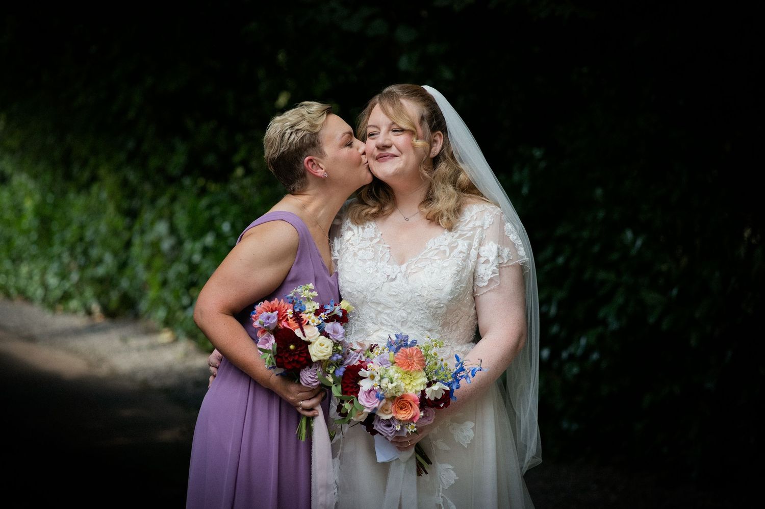 Bridesmaid in lavender dress kissing bride on cheek, both holding colorful flower bouquets outdoors.