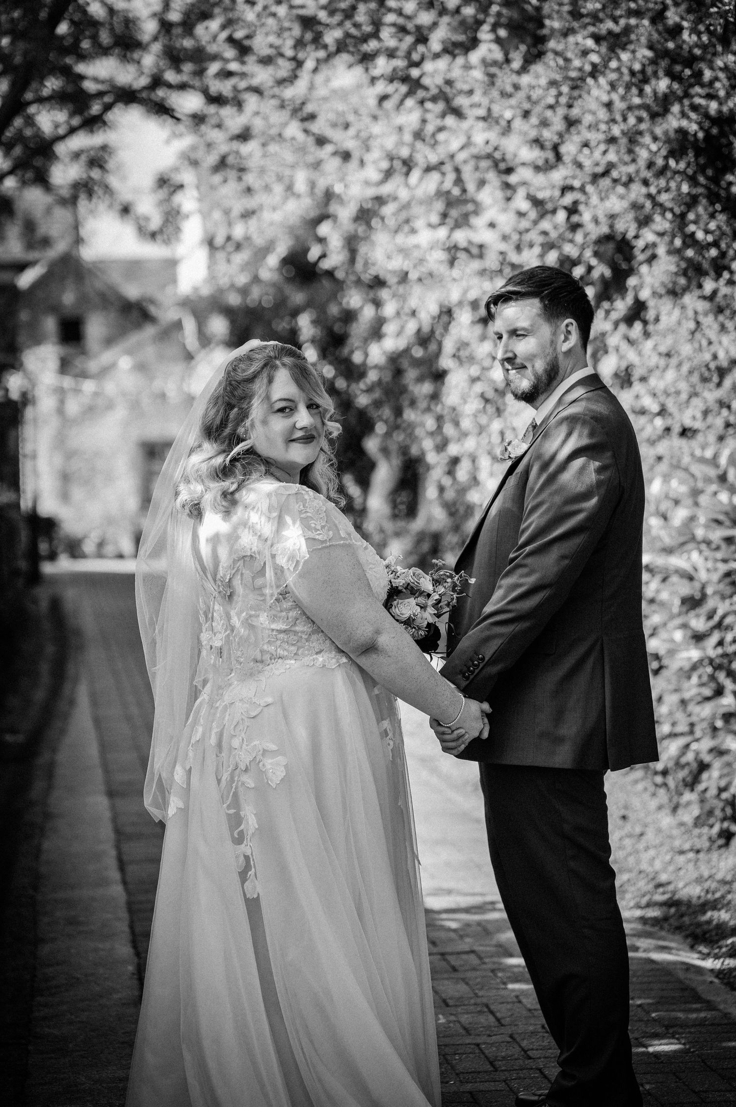 Black and white portrait of a bride and groom holding hands on a tree-lined path.