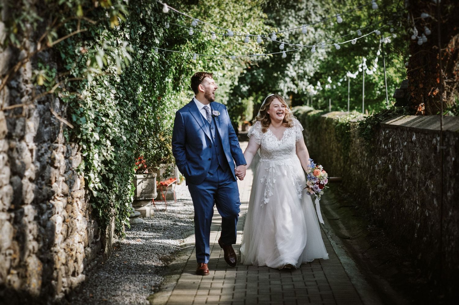Bride and groom walk hand in hand along ivy-covered stone wall pathway.