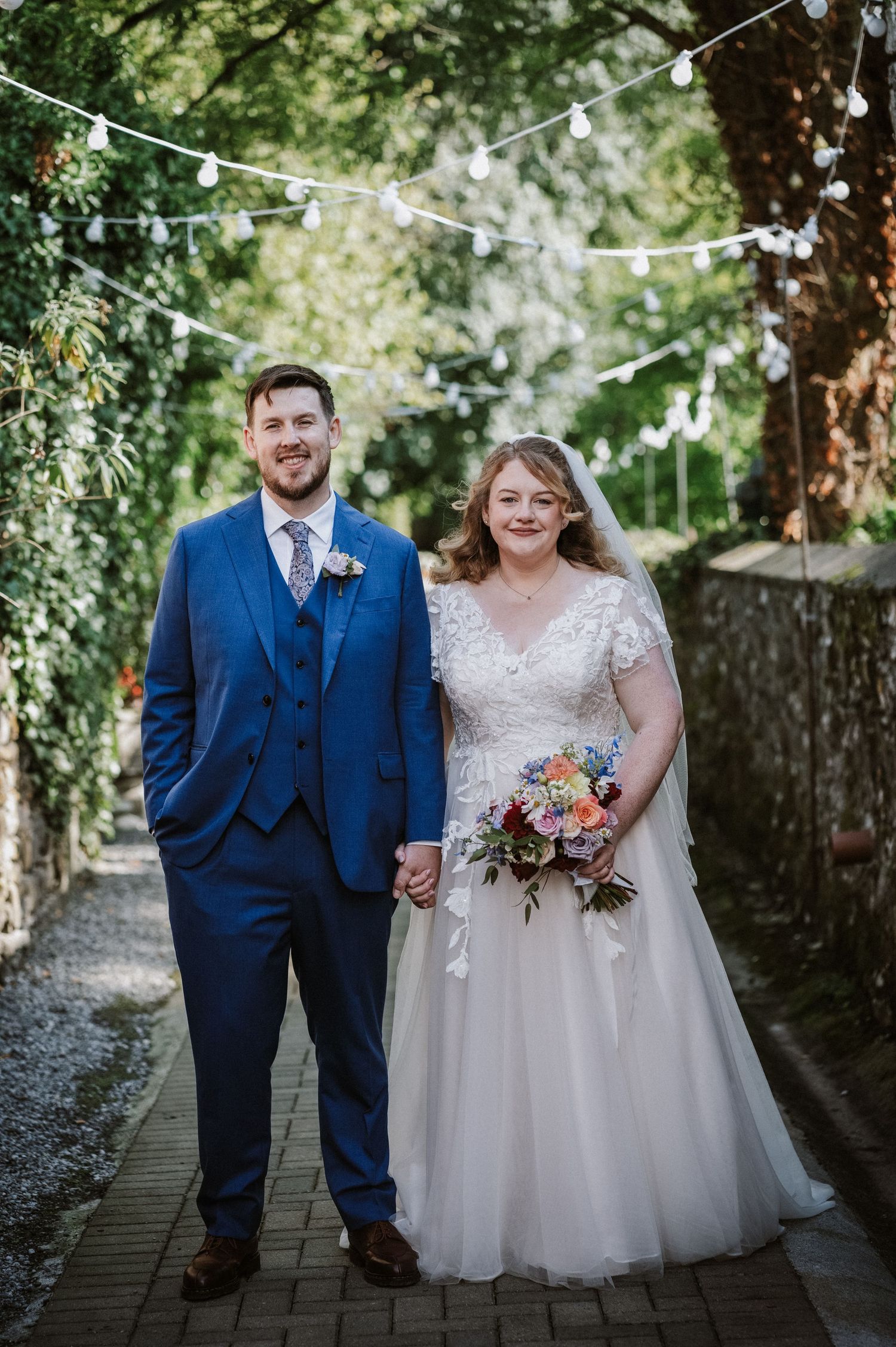 Bride and groom pose under festoon lights on a tree-lined garden pathway.