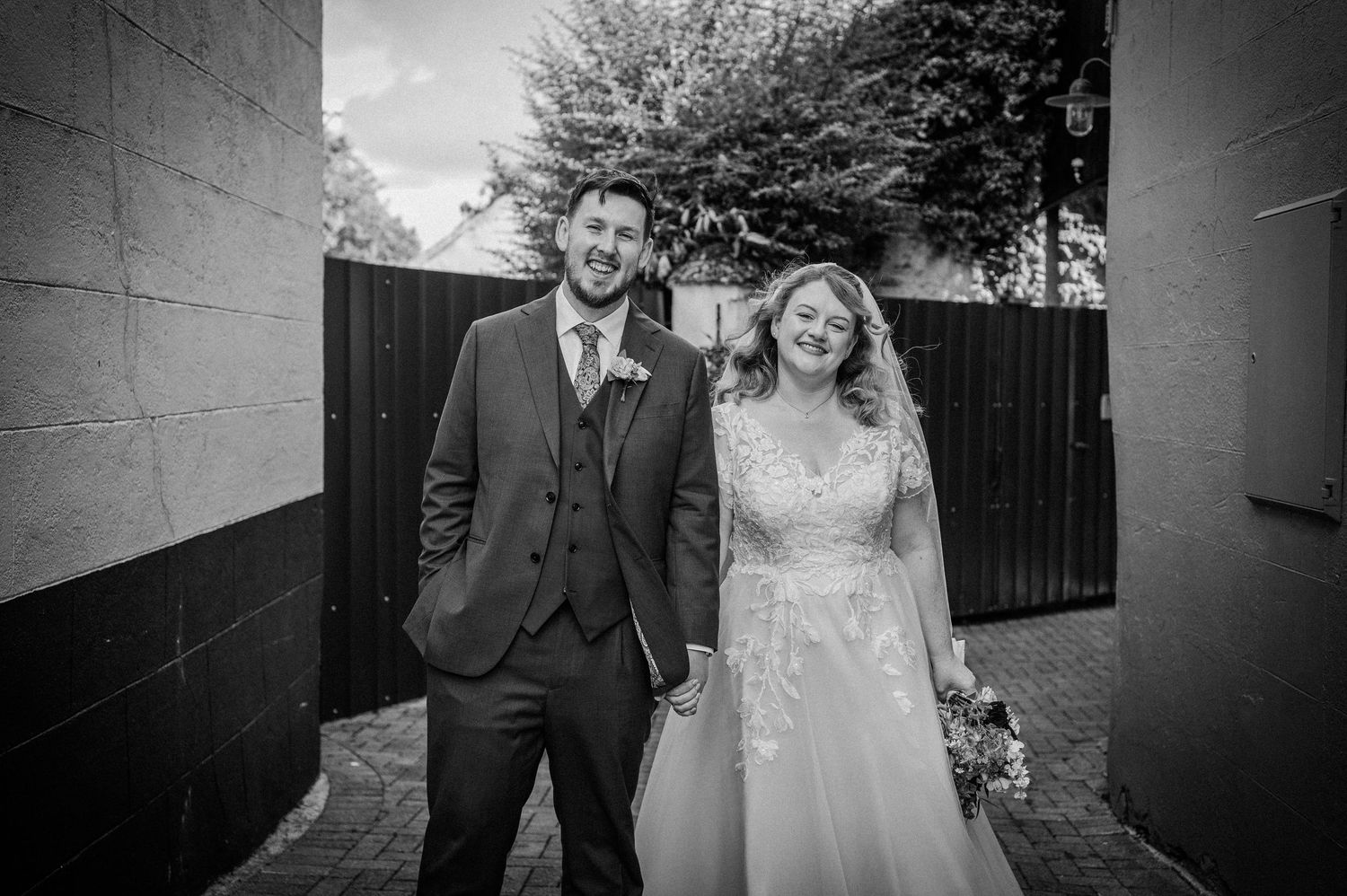 Black and white photo of smiling bride and groom holding hands between two walls outdoors.