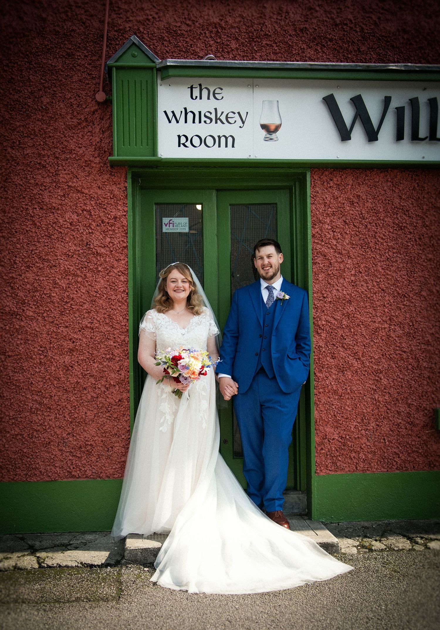 Bride and groom holding hands outside The Whiskey Room pub, bride in white gown with bouquet, groom in blue suit.