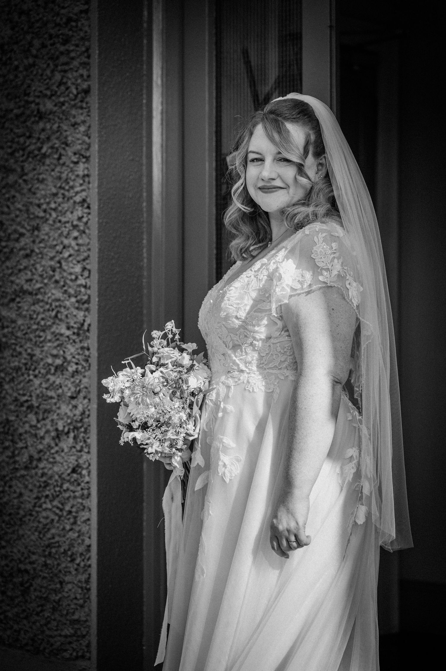 Black and white portrait of bride in lace gown and veil holding bouquet beside a textured wall.