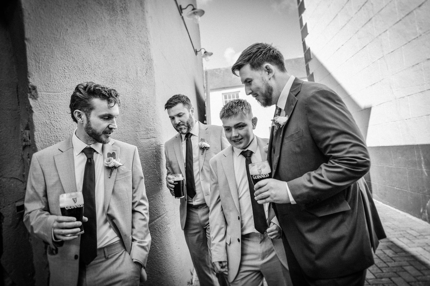 Black and white photo of four groomsmen in suits laughing and socializing outdoors at a wedding.