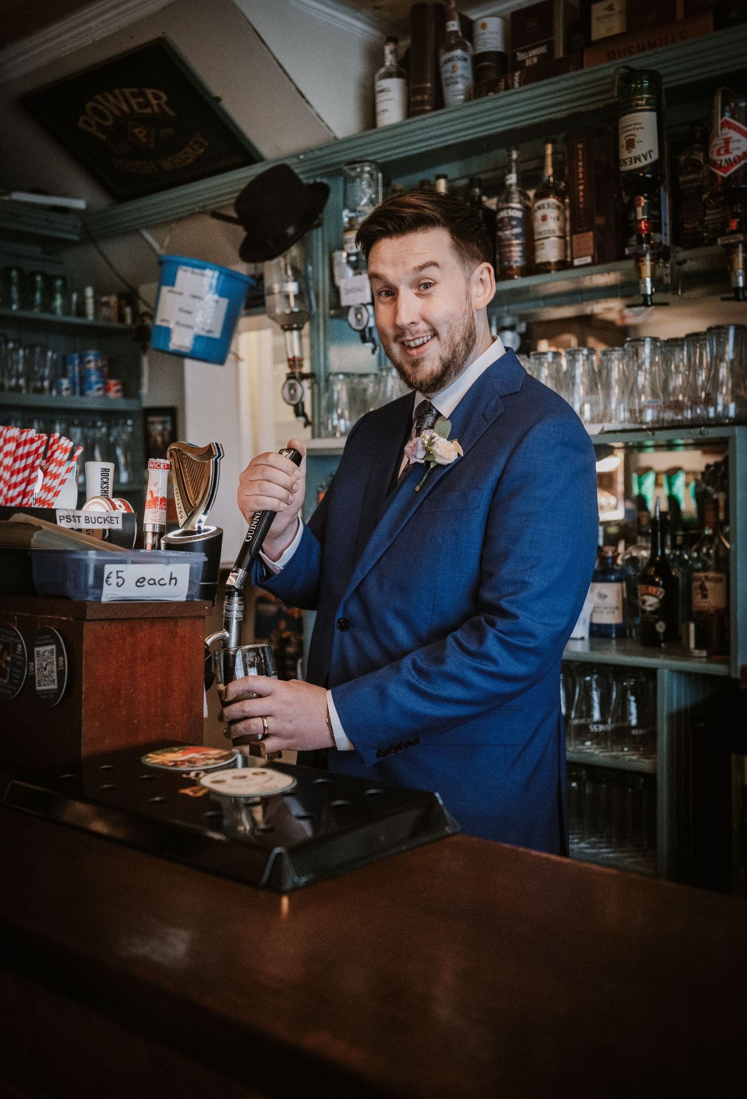Smiling groom in blue suit pours a pint of Guinness at a pub bar during wedding reception.