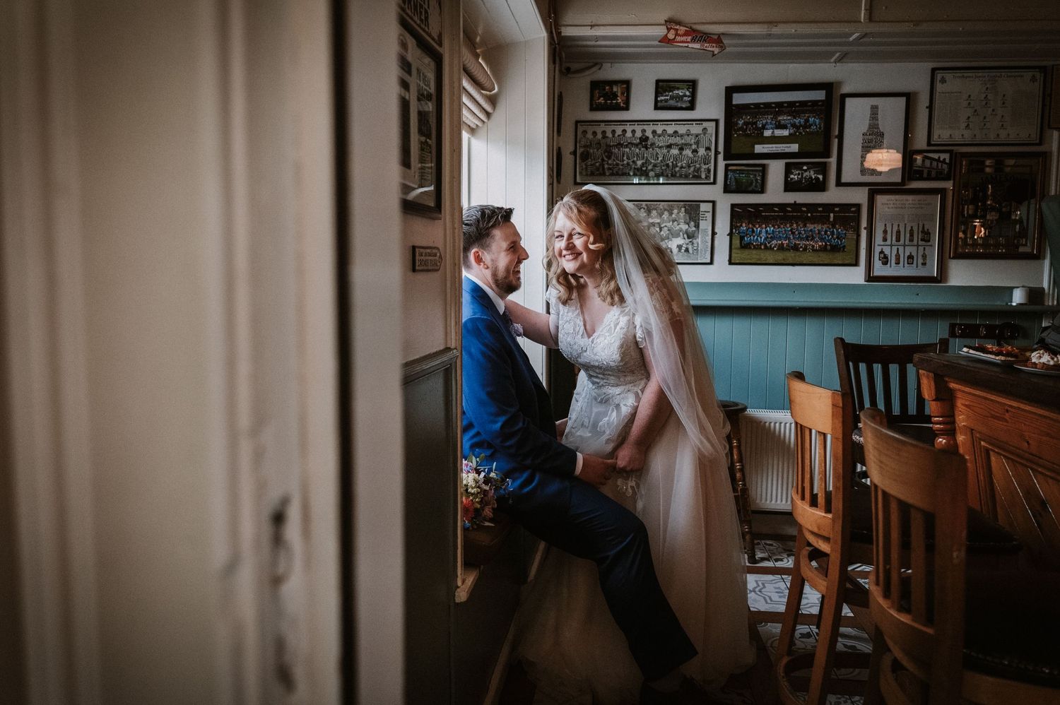 Bride and groom share intimate moment seated by window in rustic pub decorated with framed photos.