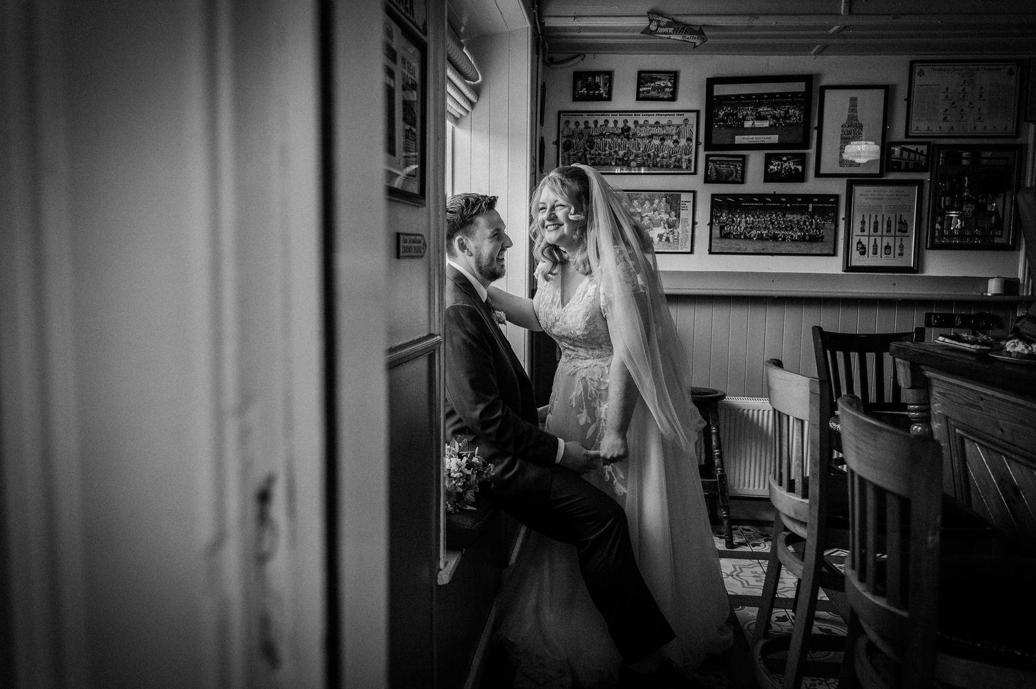 Black and white wedding photo of bride and groom sharing an intimate moment by a window in a rustic room.